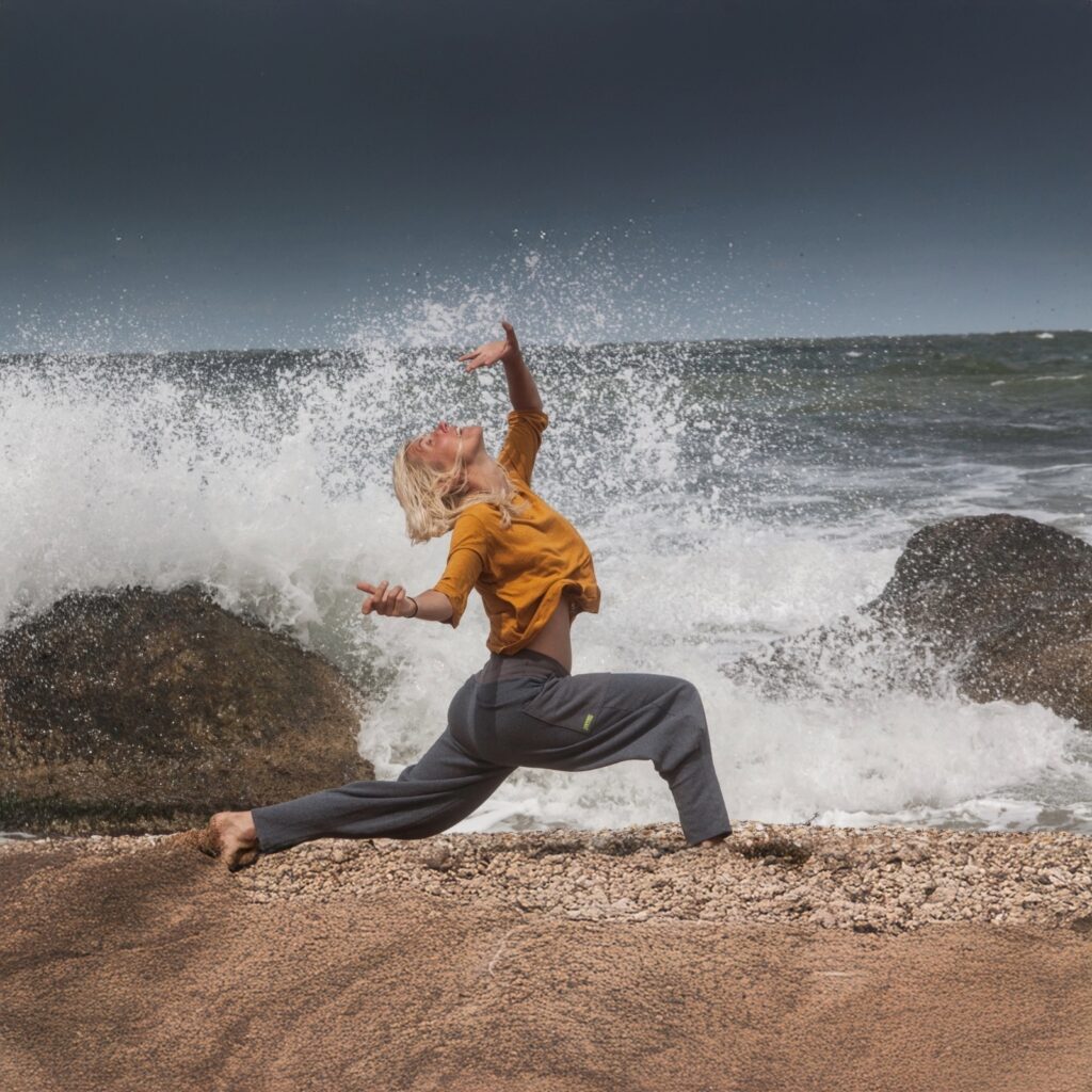 Woman practicing expressive somatic movement by the ocean with waves crashing behind her, symbolizing emotional release and healing.
