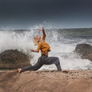 Woman practicing expressive somatic movement by the ocean with waves crashing behind her, symbolizing emotional release and healing.