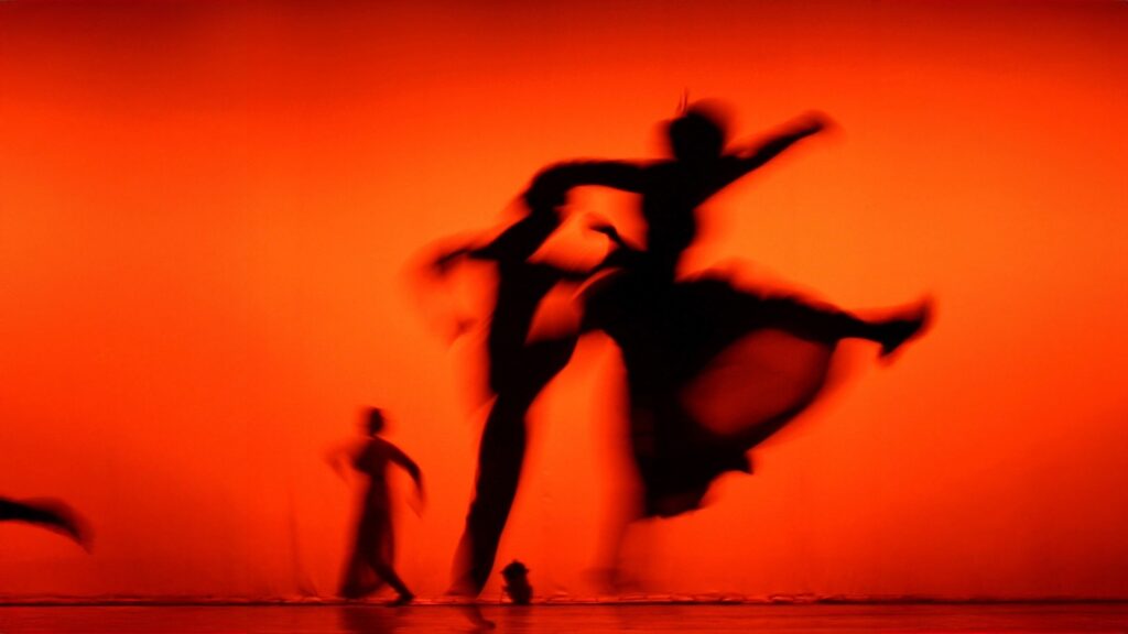 Silhouette of dancer leaping with flowing fabric against vibrant orange background representing emotional release and somatic movement therapy
