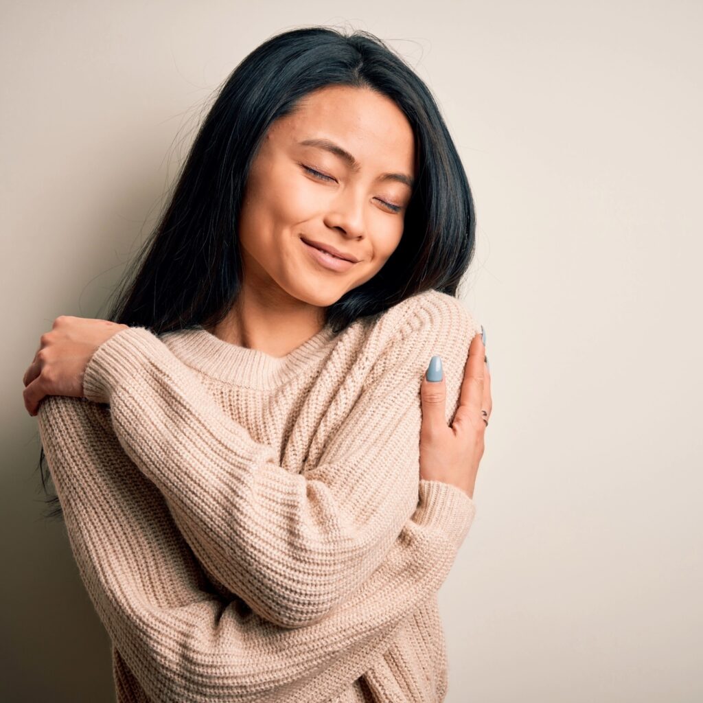 Woman with dark hair hugging herself with eyes closed, practising self-soothing and nervous system regulation through gentle self-embrace